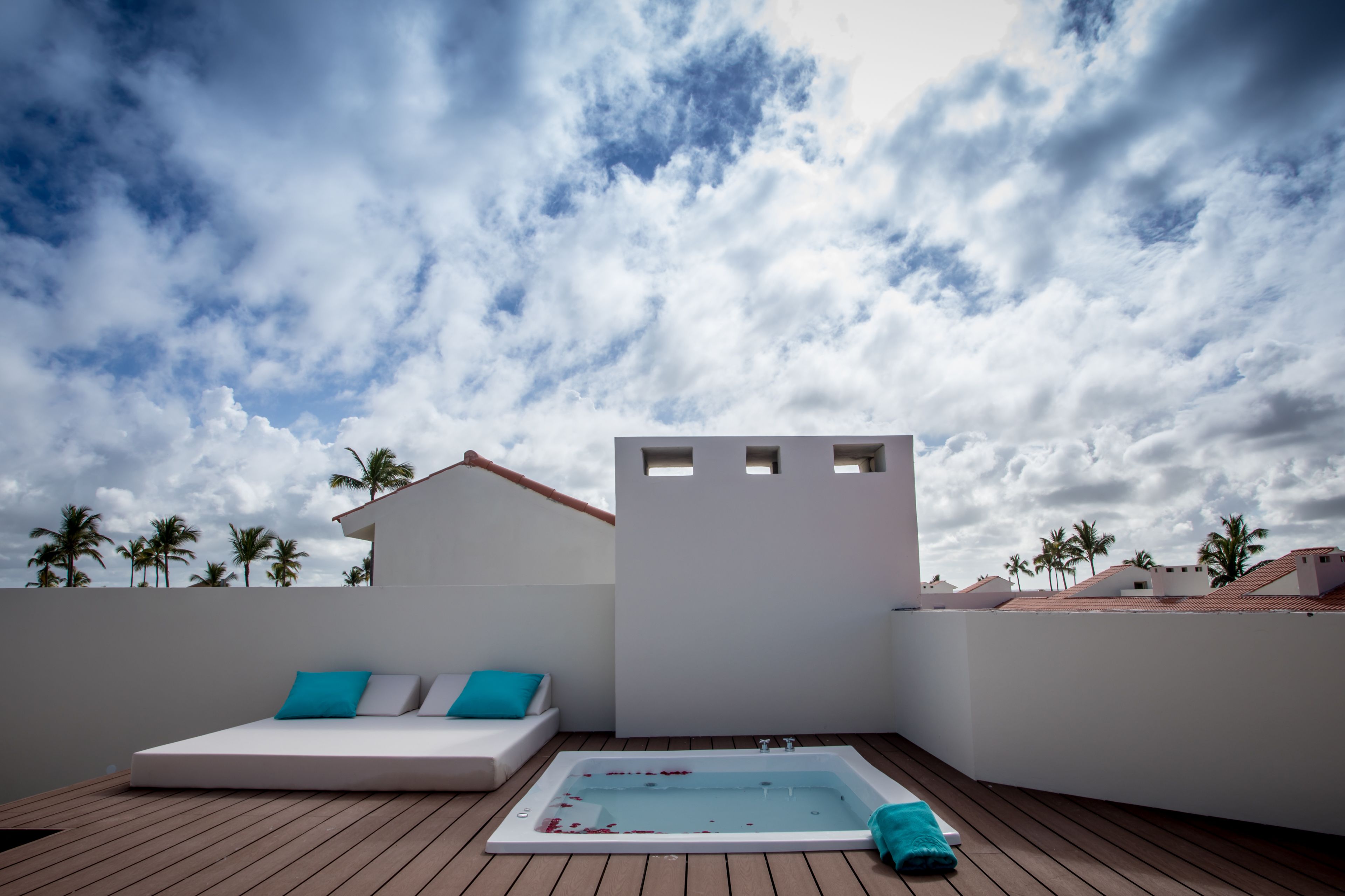 Une terrasse sur le toit avec une piscine, un espace détente avec un canapé et des coussins, et une vue sur les palmiers et les bâtiments sous un ciel nuageux.
