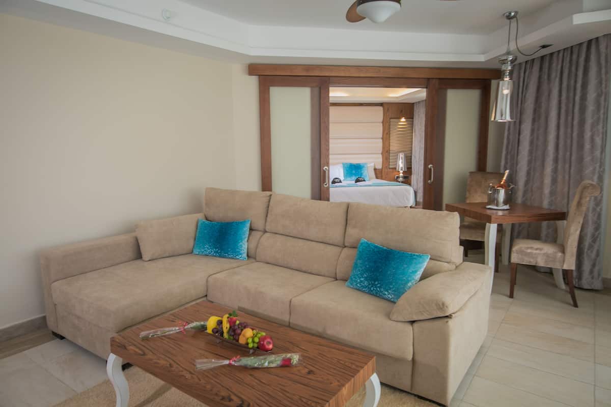 A modern living room with a beige sectional sofa, a wooden coffee table with fruit, and a view into the bedroom through a sliding door.