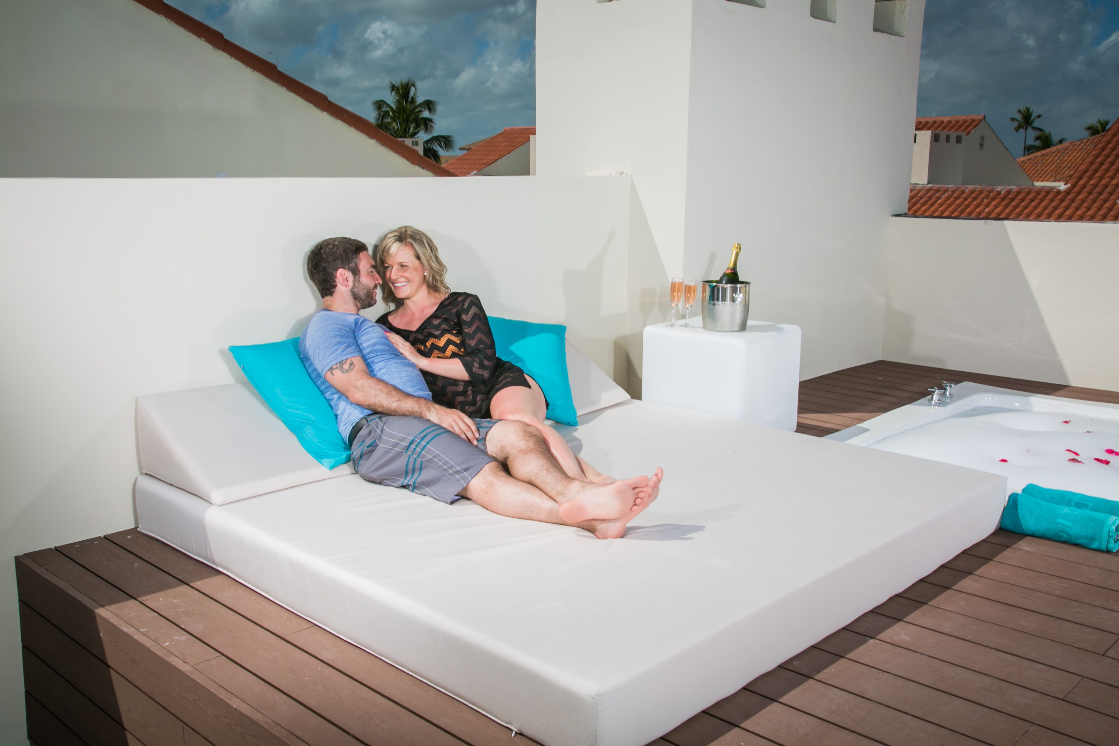 A couple sitting on a white sofa with a blue pillow, a small table with a bottle and glasses, and a wooden deck.