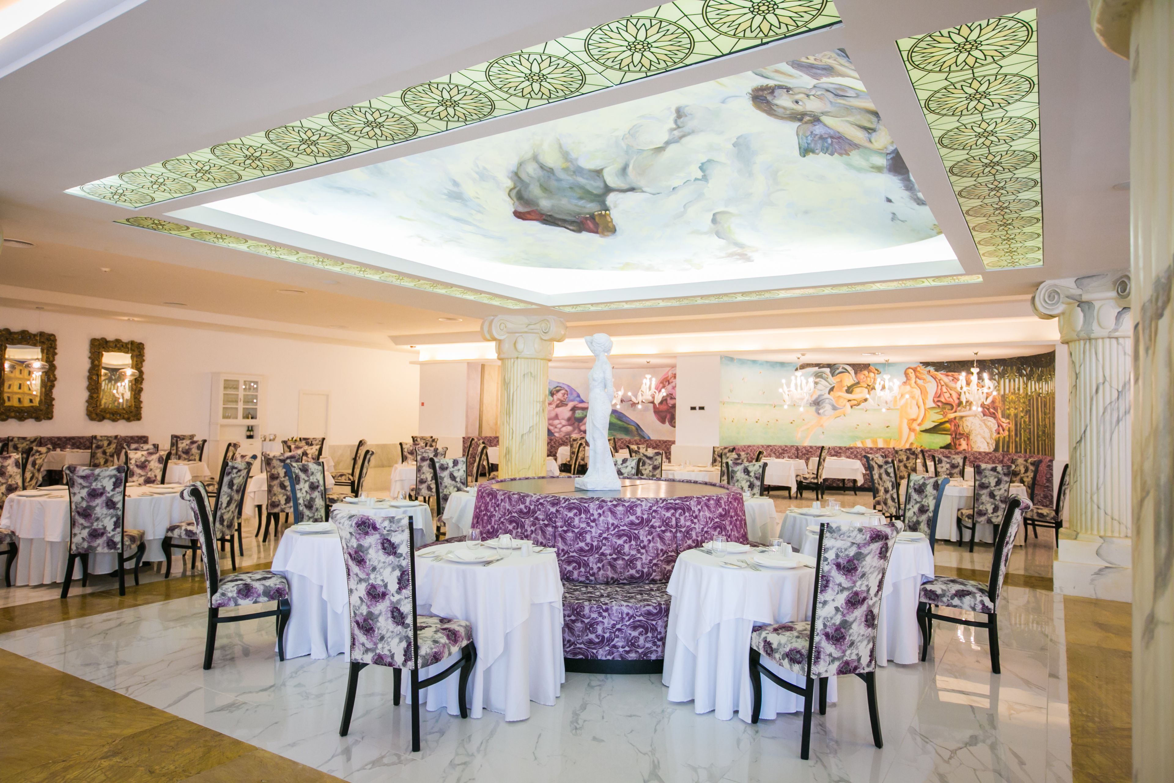 Un vaste espace dédié à la salle à manger, avec des tables rondes et des chaises à motifs floraux, un coin salon central agrémenté d’un canapé violet, et un plafond orné d’un motif floral.