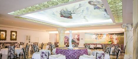 Un vaste espace dédié à la salle à manger, avec des tables rondes et des chaises à motifs floraux, un coin salon central agrémenté d’un canapé violet, et un plafond orné d’un motif floral.