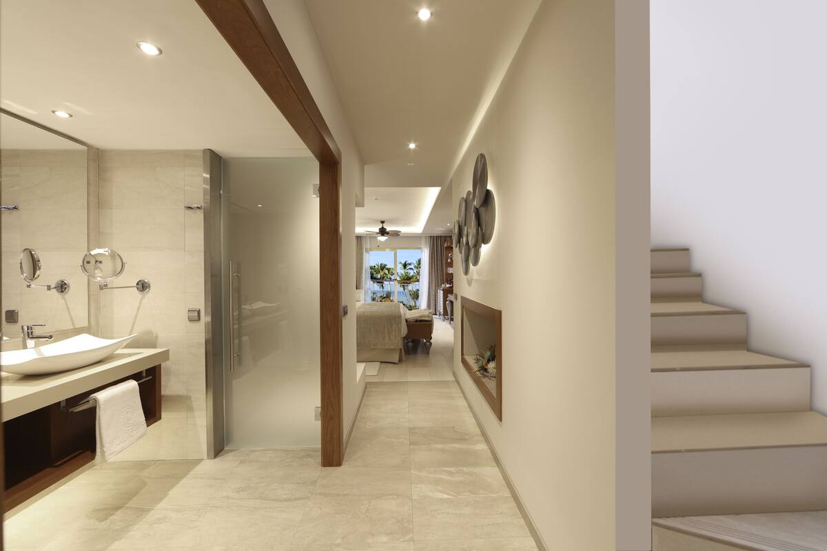 A modern bathroom with a large sink, mirror, and shower. A hallway leads to a bedroom with a bed and a view of the outdoors.