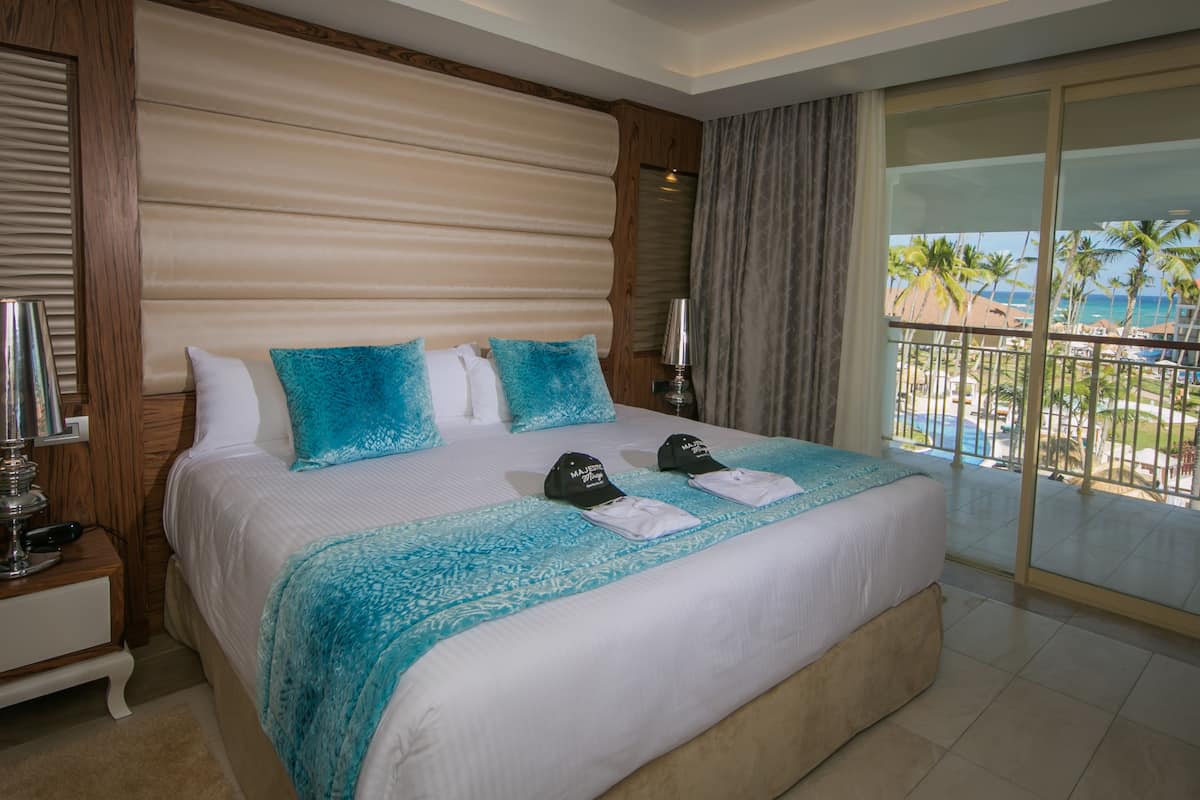 A hotel room with a large bed, a nightstand, a lamp, and a view of a beach and palm trees.