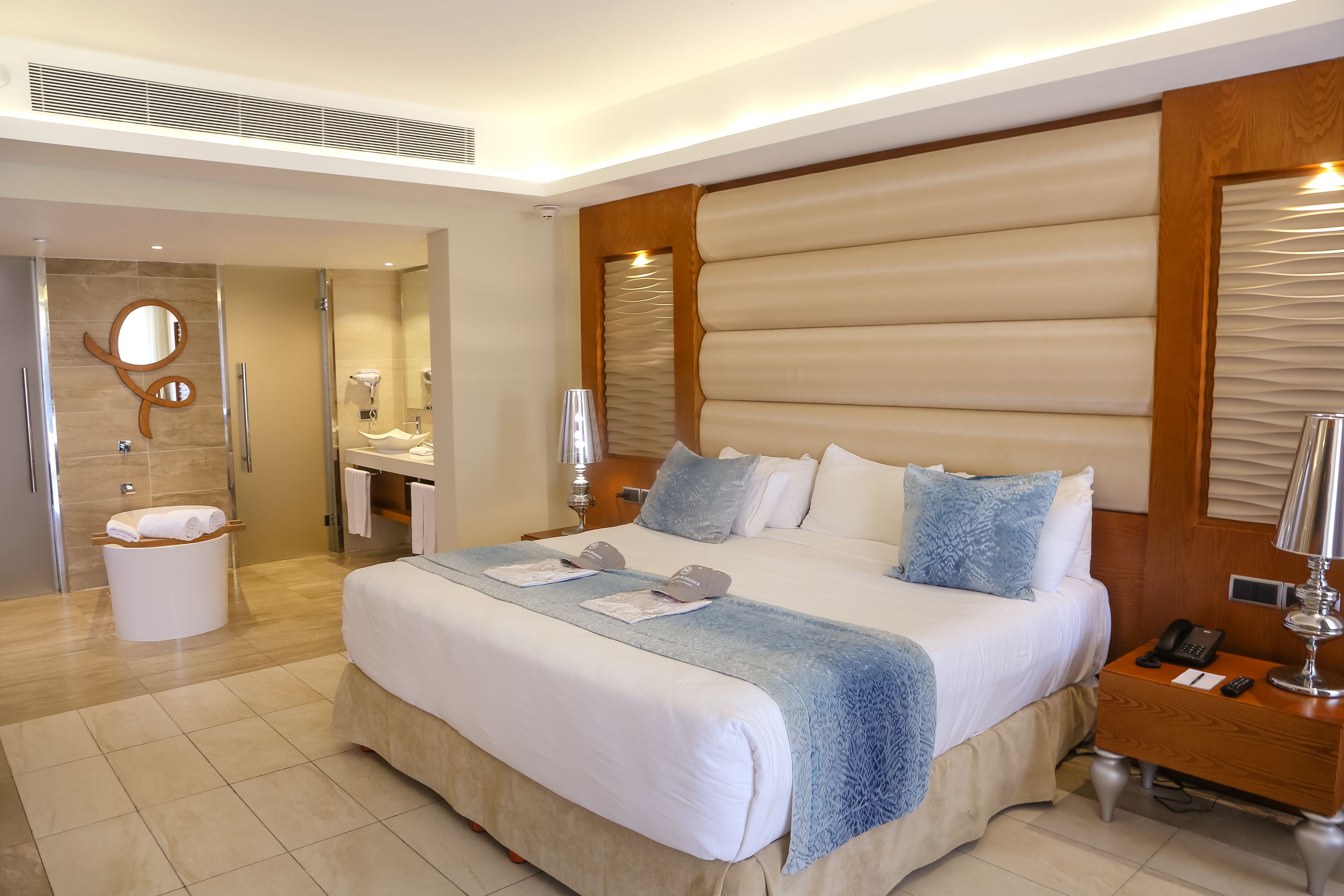 Een moderne hotelkamer met een groot bed, een houten hoofdbord, een nachtkastje met een lamp, en een badkamer op de achtergrond.