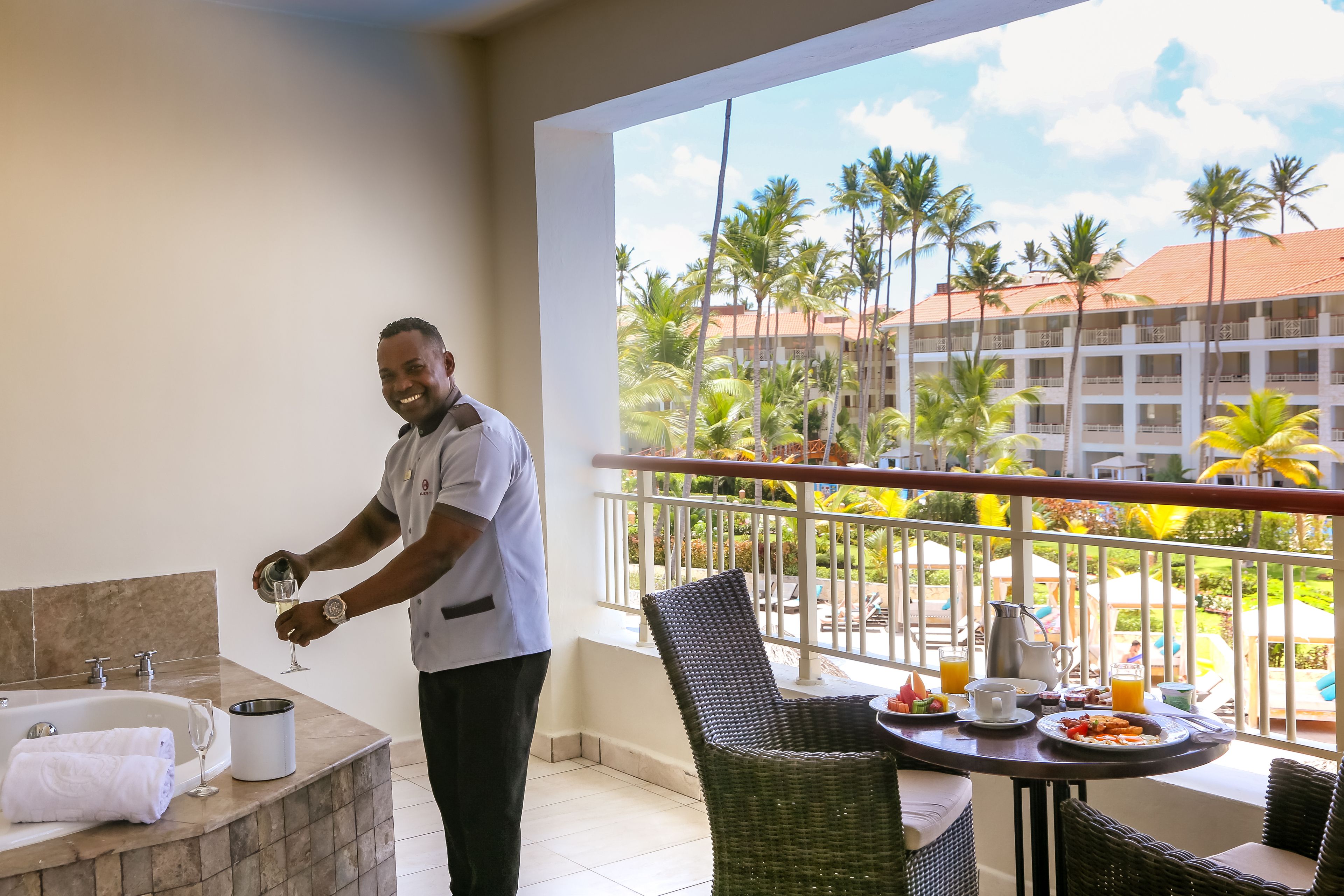 A hotel room with a balcony, a bathtub, a dining table with breakfast, and a view of palm trees and a building.