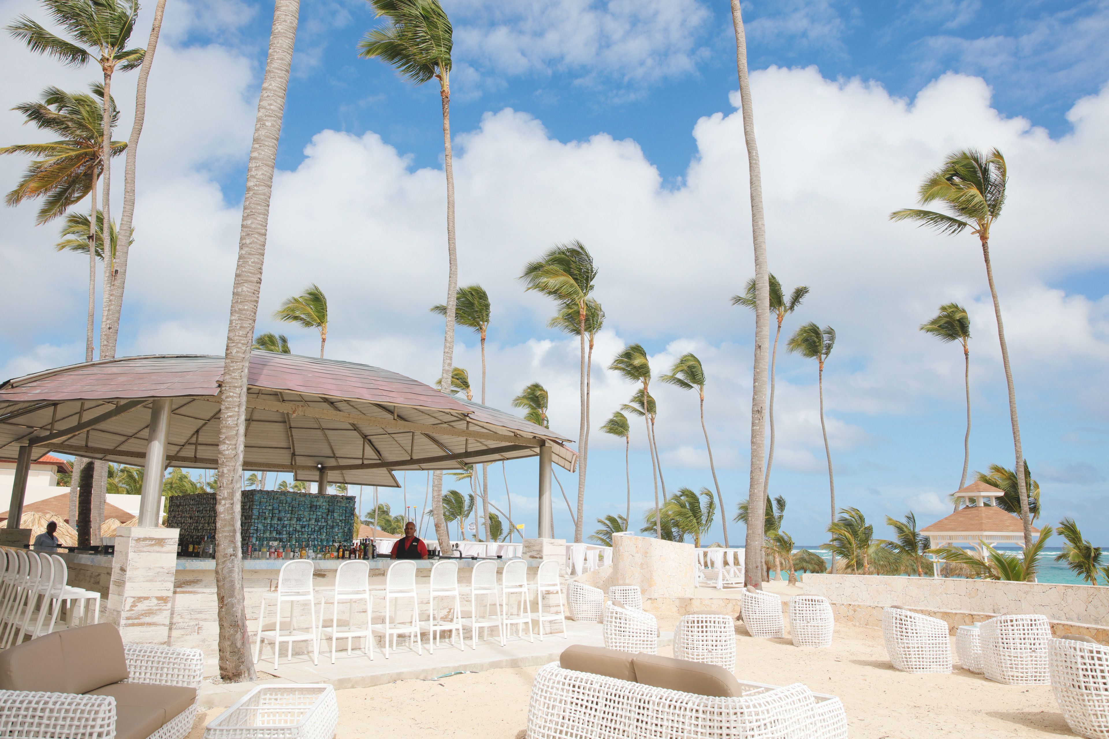 Un espace aménagé près de la plage, avec des meubles en osier blancs, un pavillon couvert et des palmiers sous un ciel dégagé.