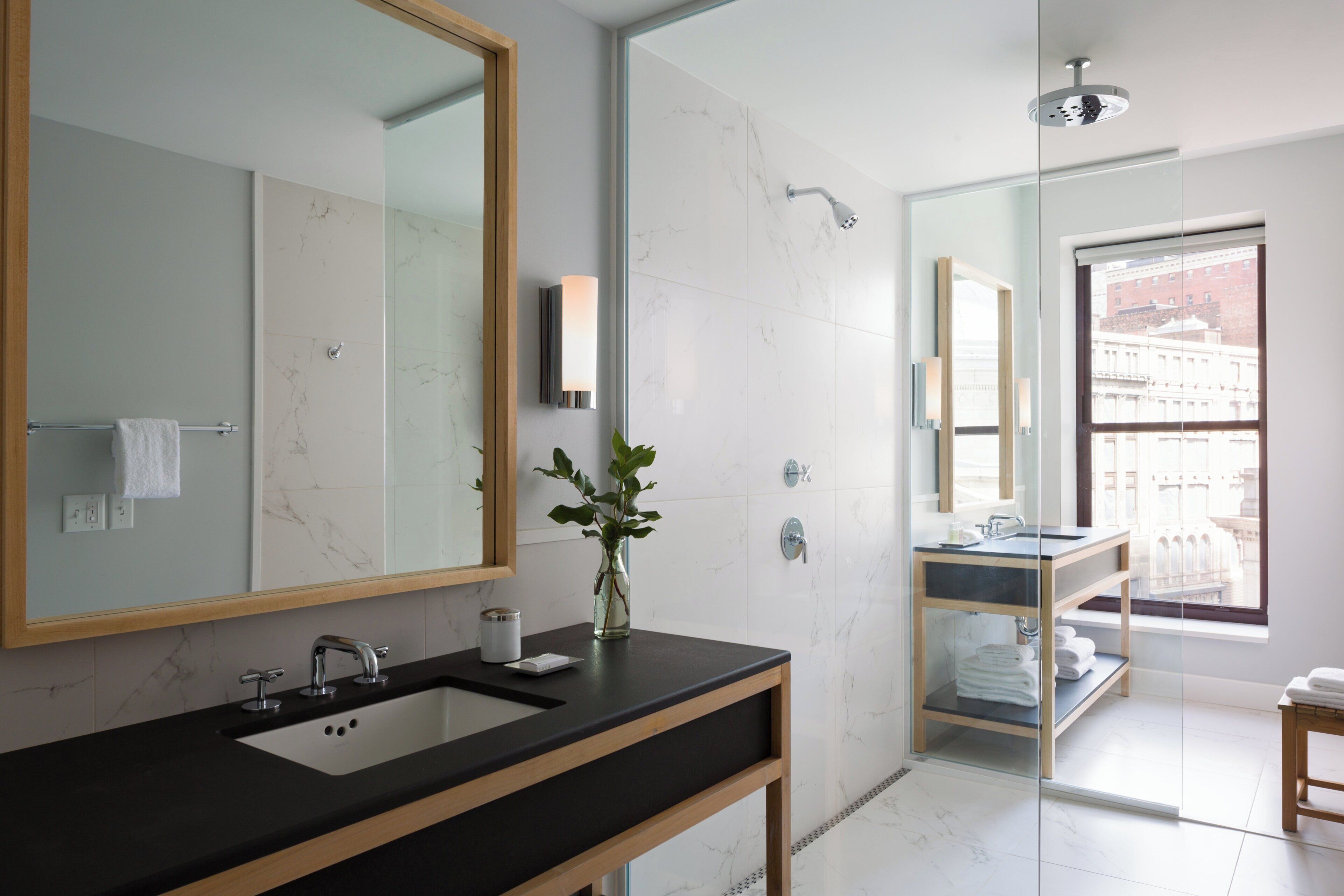 A modern bathroom with a large mirror, a sink, a shower, and a view of a building through a window.