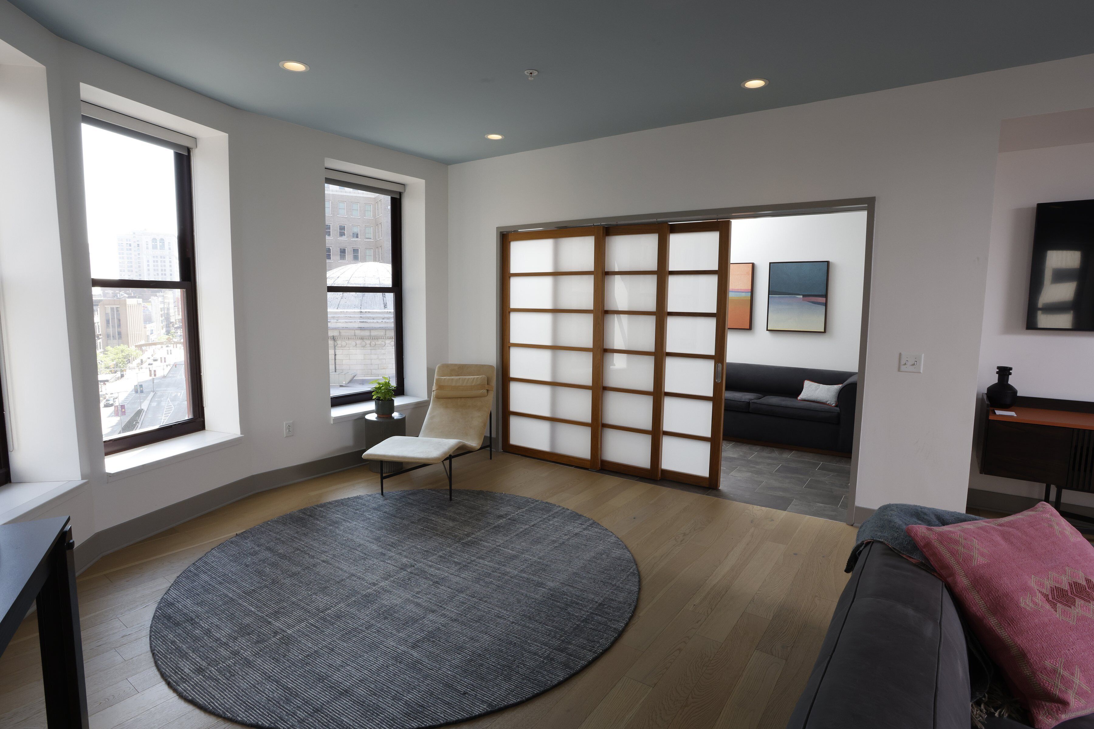A modern living room with a large window, a sliding glass door, a round rug, a sofa, and a small table.