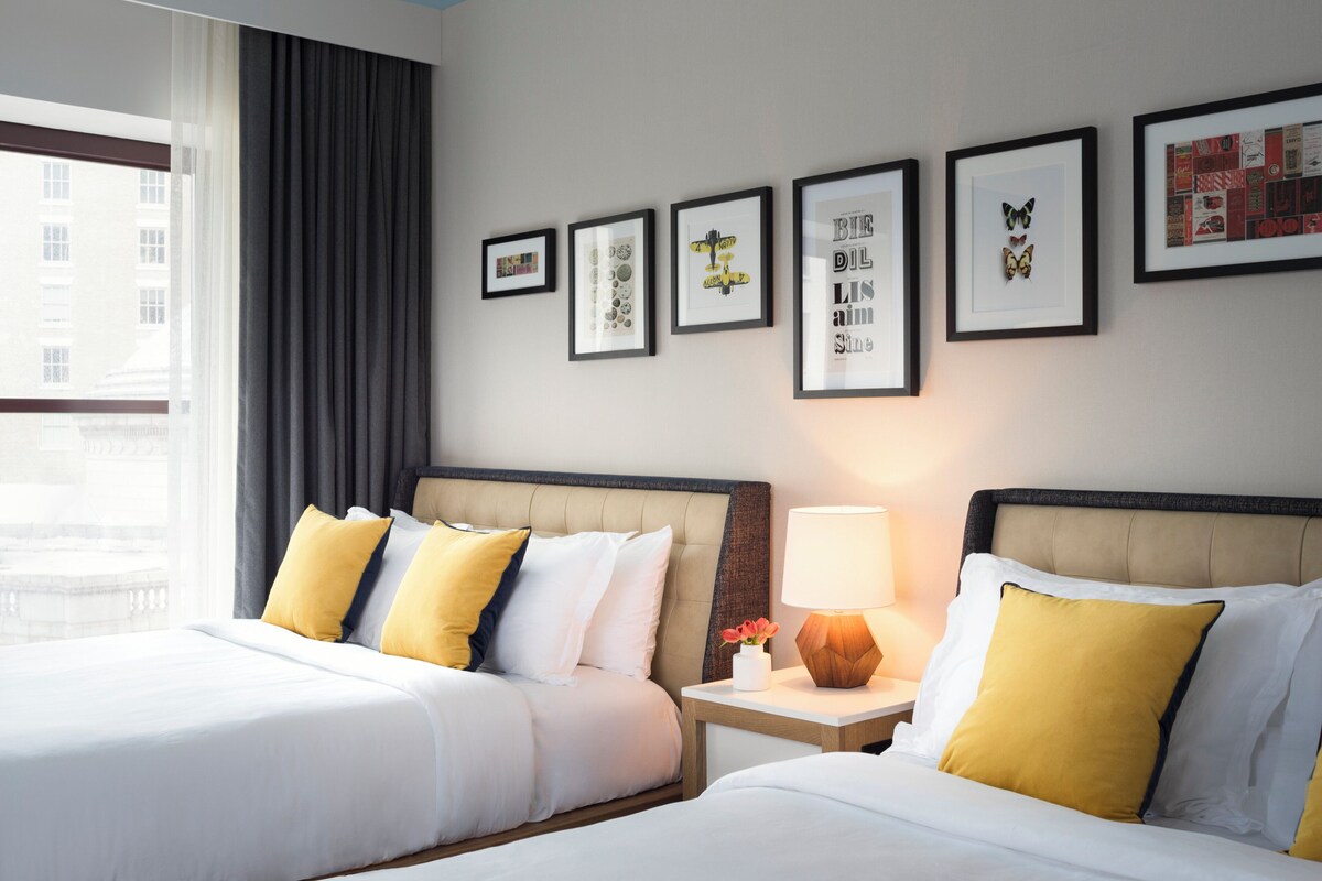Een hotelkamer met twee bedden, een nachtkastje met een lamp, en kunstwerken in een lijst aan de muur.