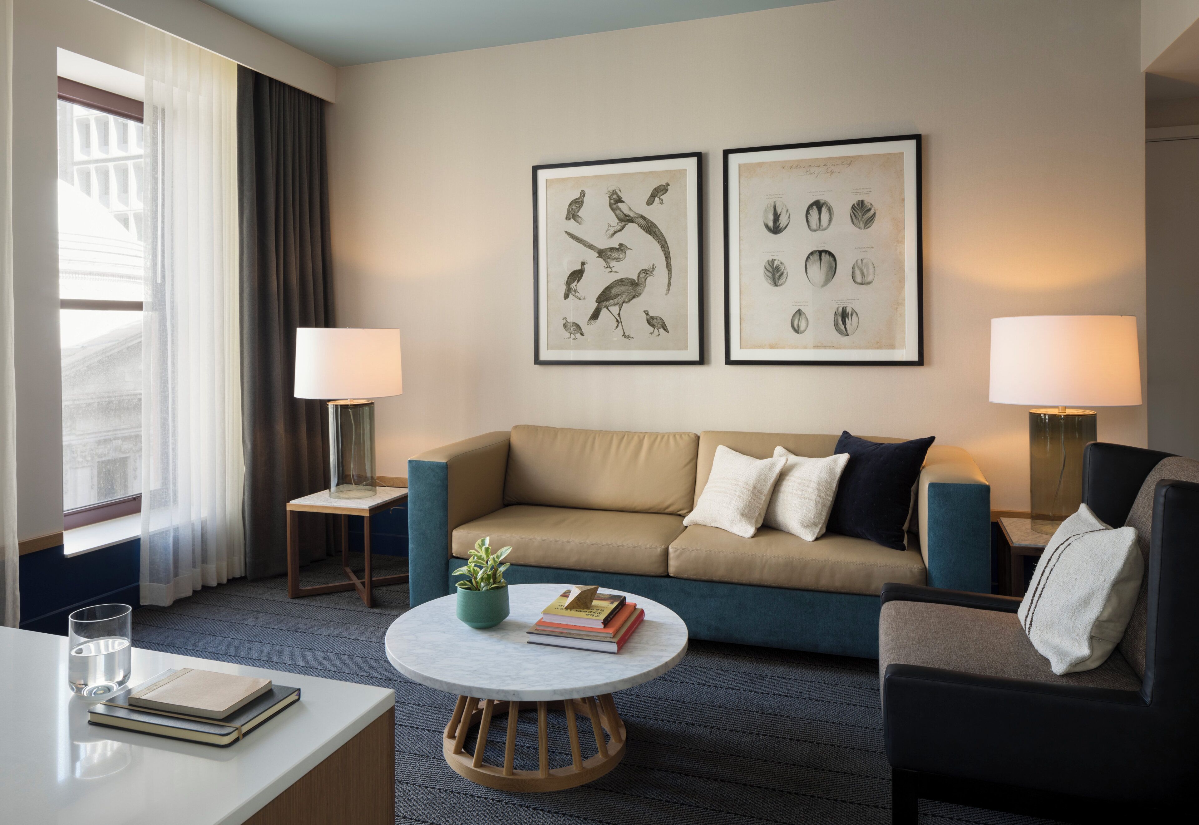 A hotel room with a beige sofa, two side tables with lamps, a round coffee table with a plant and books, and two framed botanical prints on the wall.