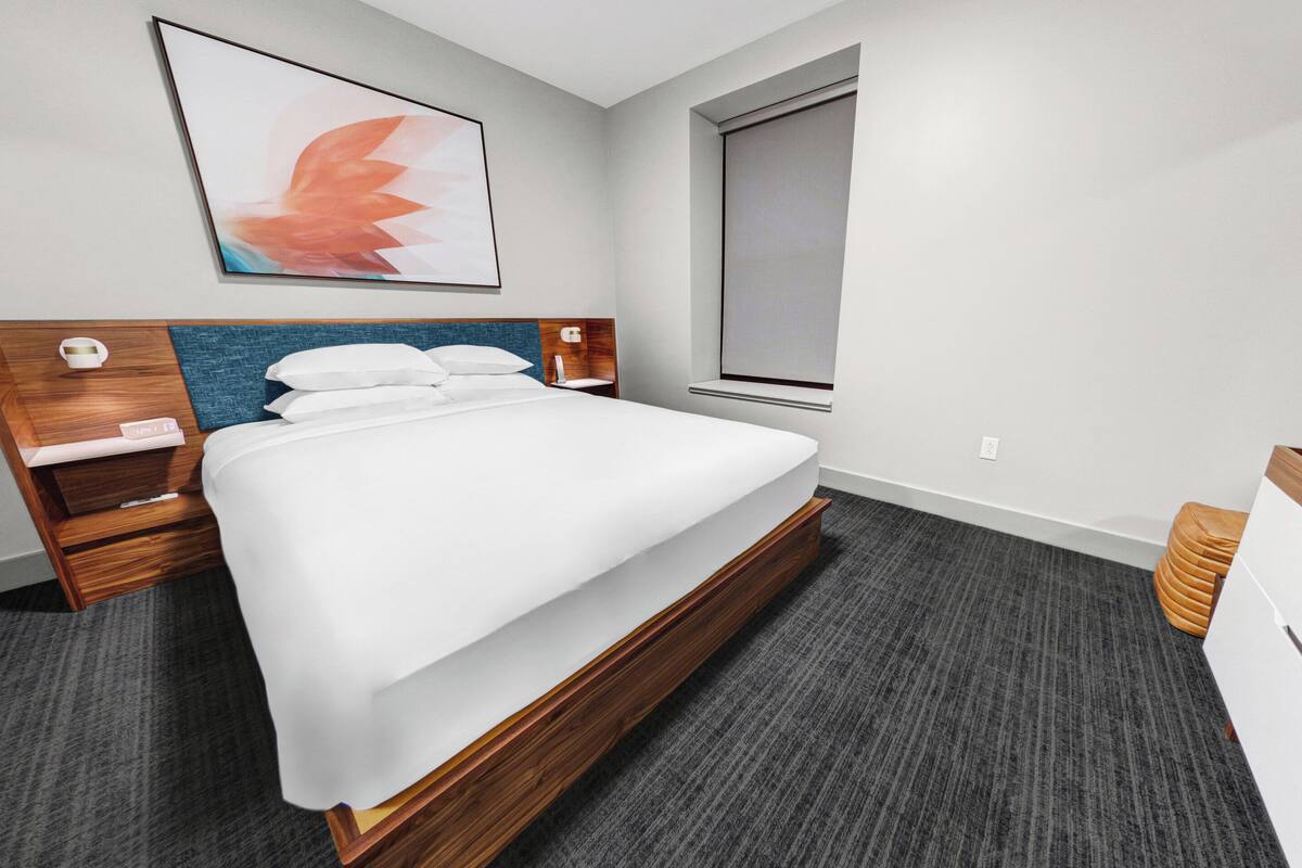 A modern hotel room with a bed, wooden headboard, a nightstand, a desk, and a window.