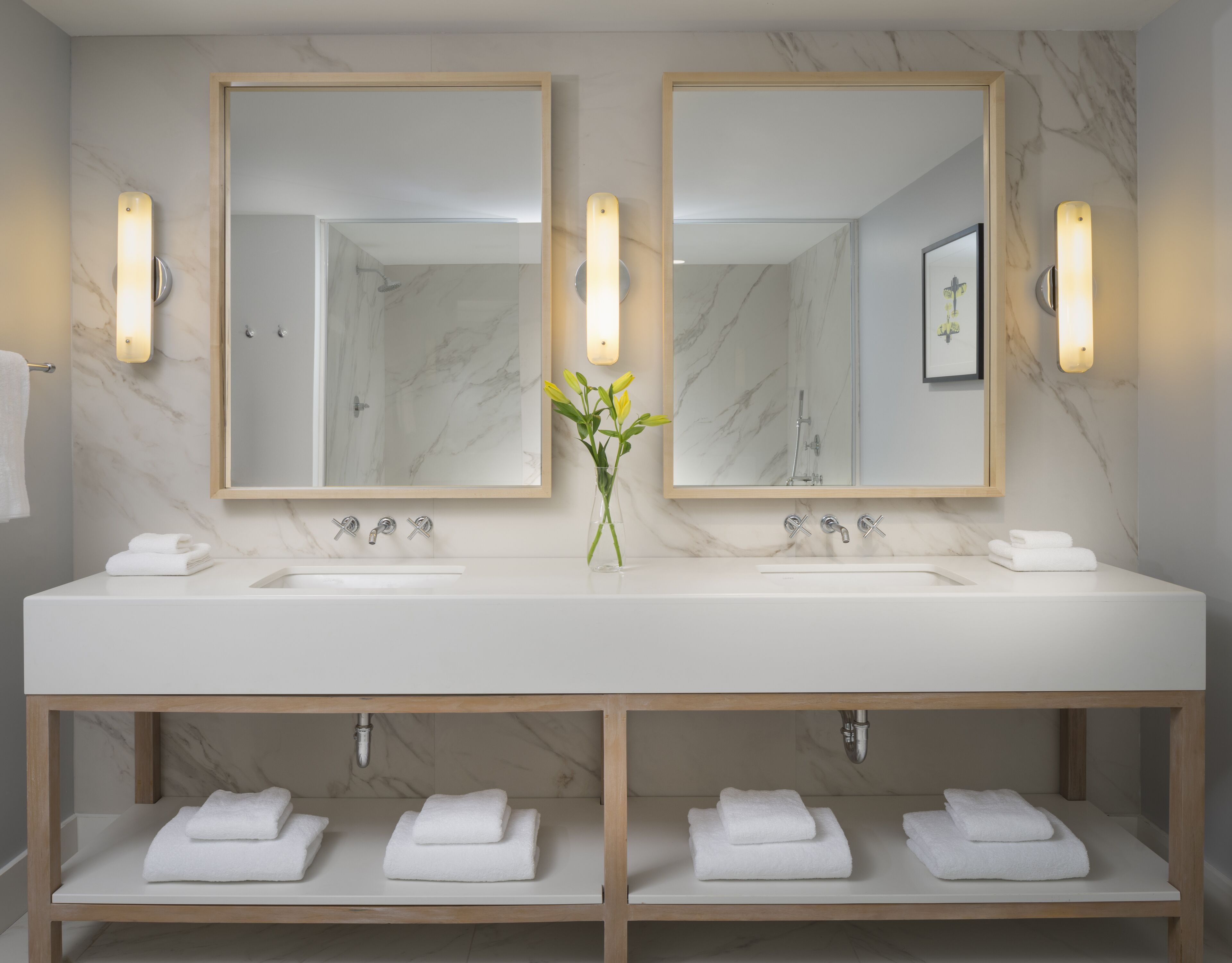 Un baño moderno con dos lavamanos, paredes de mármol y un florero con flores.