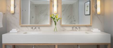 Un baño moderno con dos lavamanos, paredes de mármol y un florero con flores.