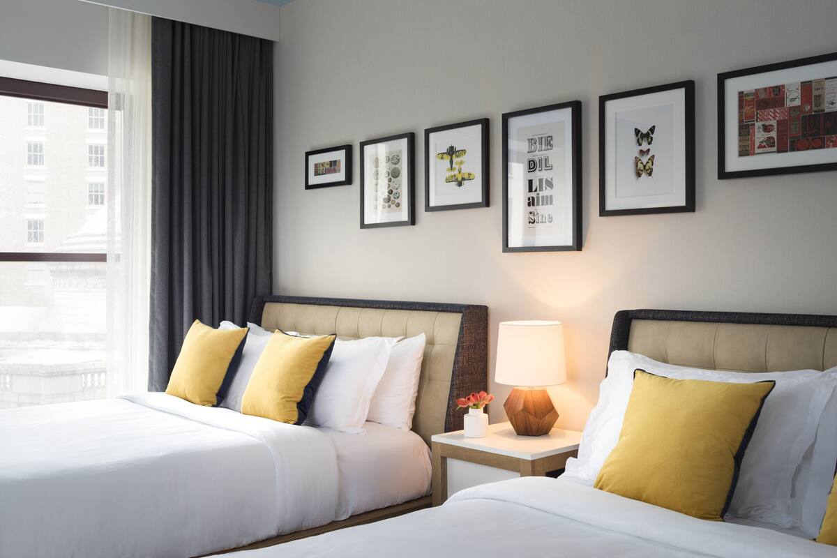 A hotel room with two beds, a nightstand with a lamp, and framed artwork on the wall.