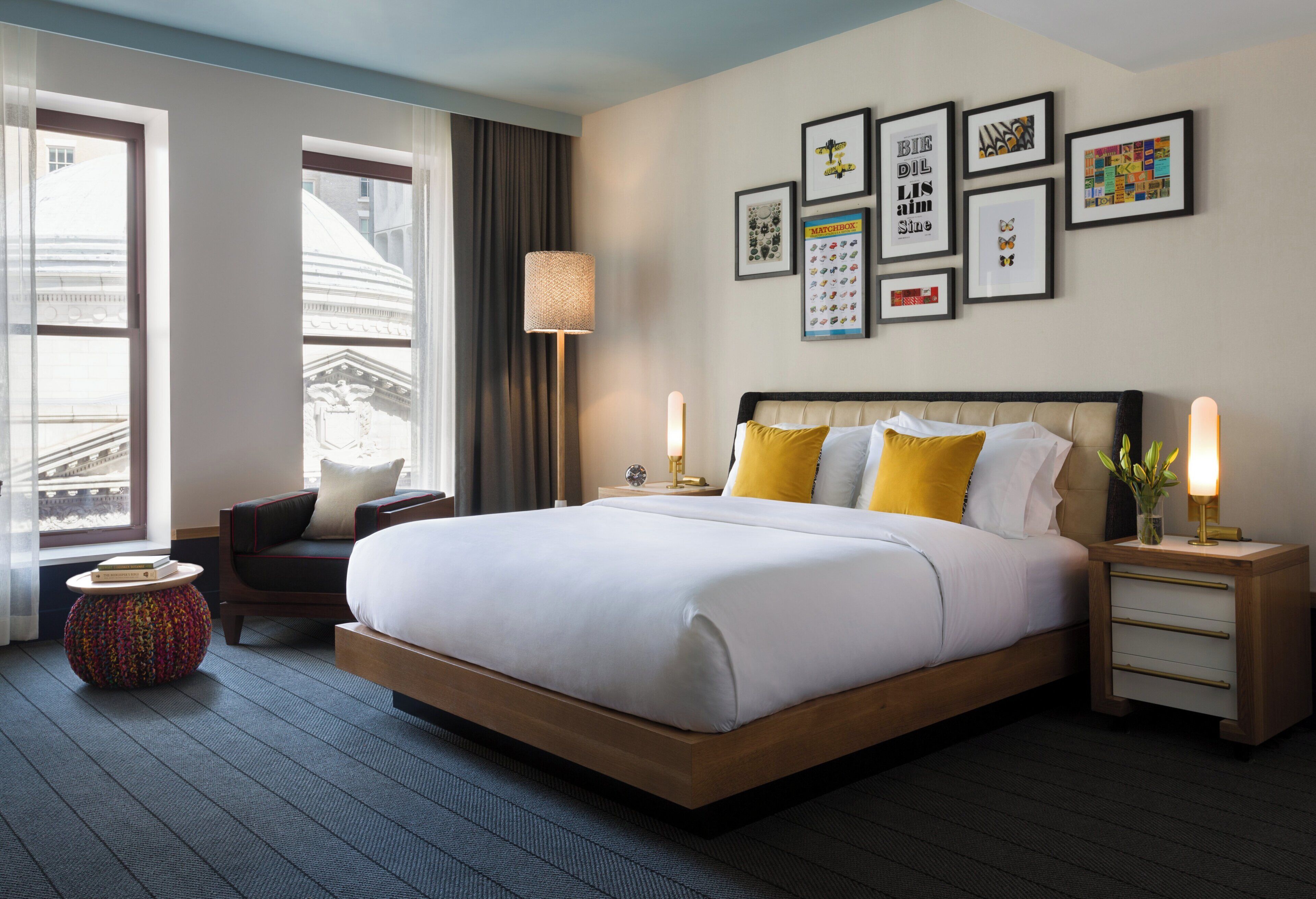 Quarto de hotel com uma cama grande, uma mesa de cabeceira, uma cadeira, um abajur e um painel com quadros.