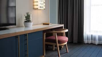 A modern hotel room with a wooden desk, a potted plant, a chair, a television, and a wall-mounted light fixture.