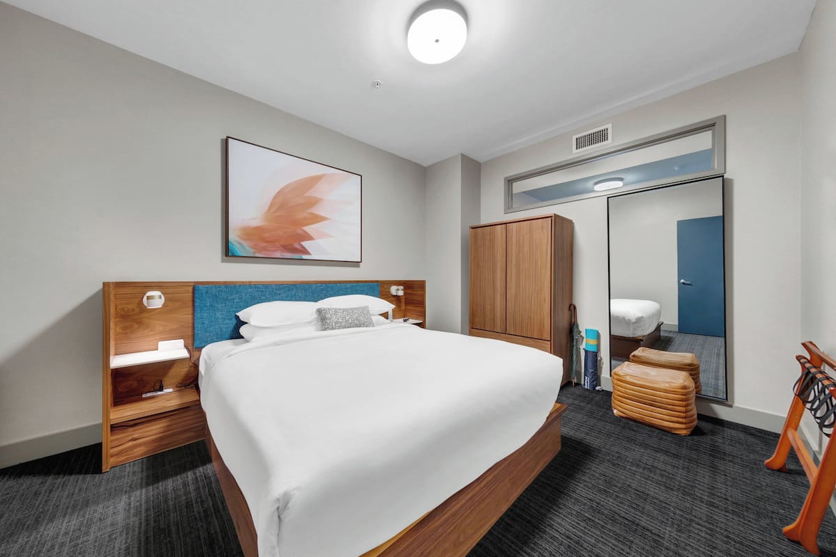 A hotel room with a large bed, a wooden headboard, a bedside table, a mirror, a wardrobe, and a TV mounted on the wall.