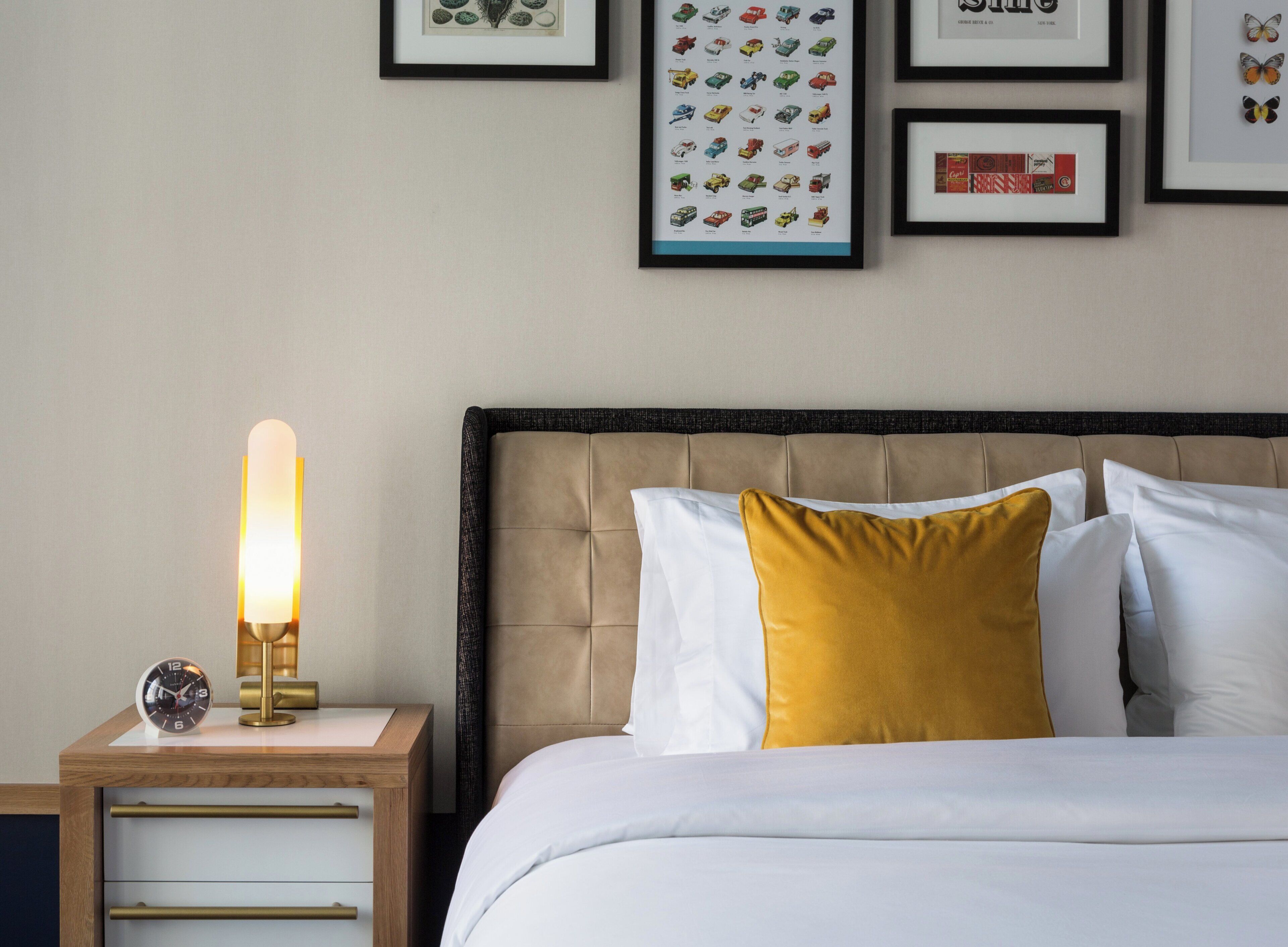 A neatly made bed with white and mustard pillows, a bedside lamp, and a clock on a wooden nightstand.