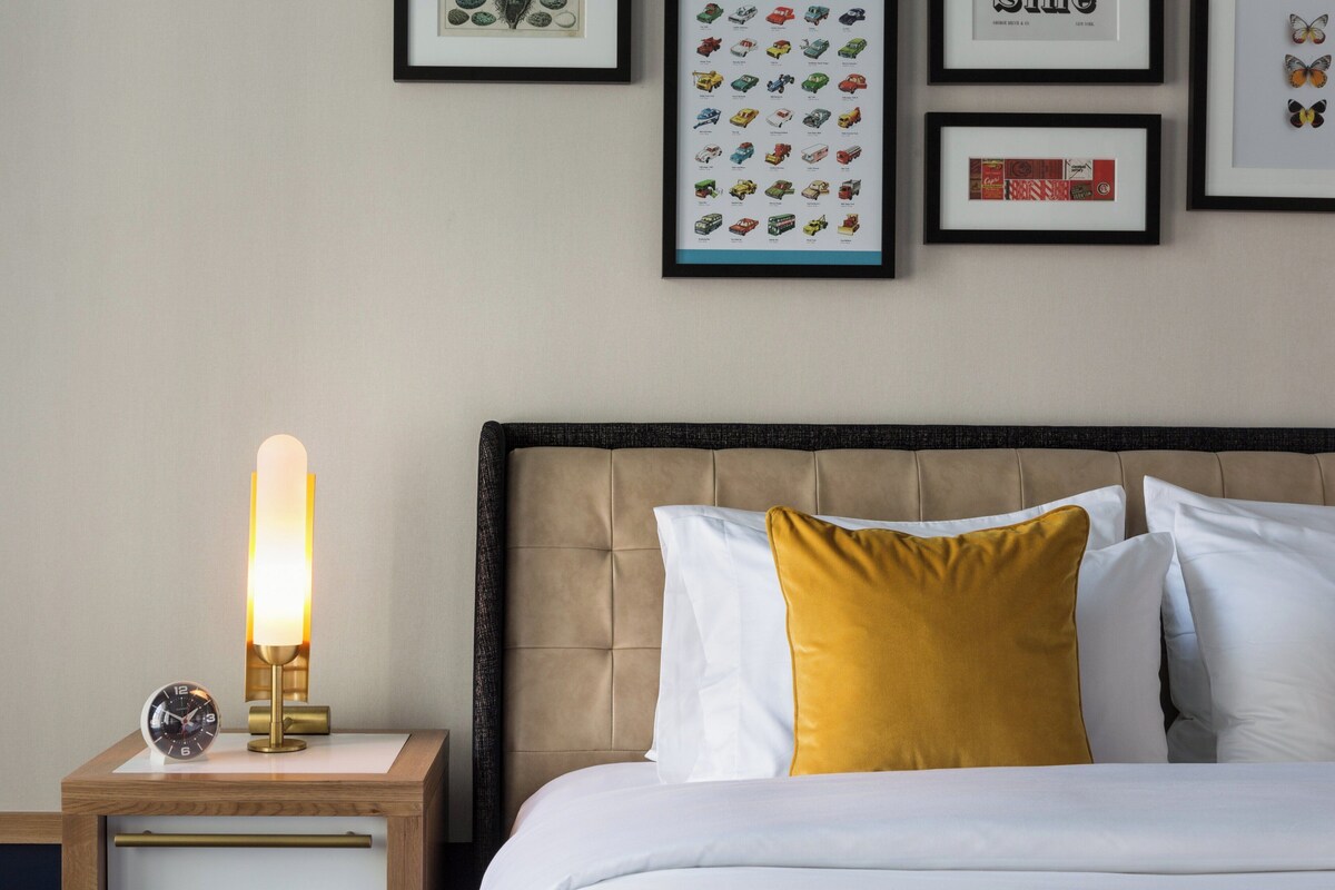 Een net opgemaakt bed met witte en mosterdkleurige kussens, een bedlampje en een klok op een houten nachtkastje.