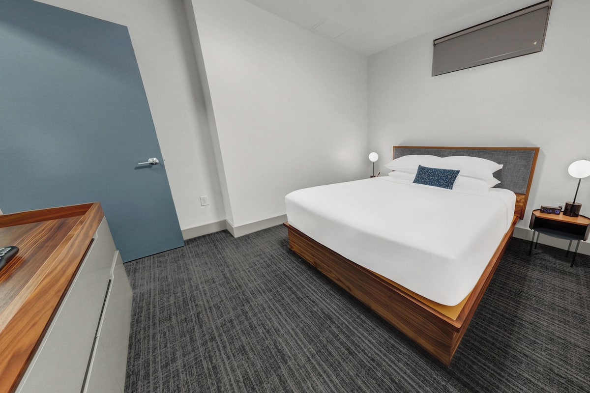 A modern hotel room with a large bed, a desk with a chair, a bedside table with a lamp, and a small window.