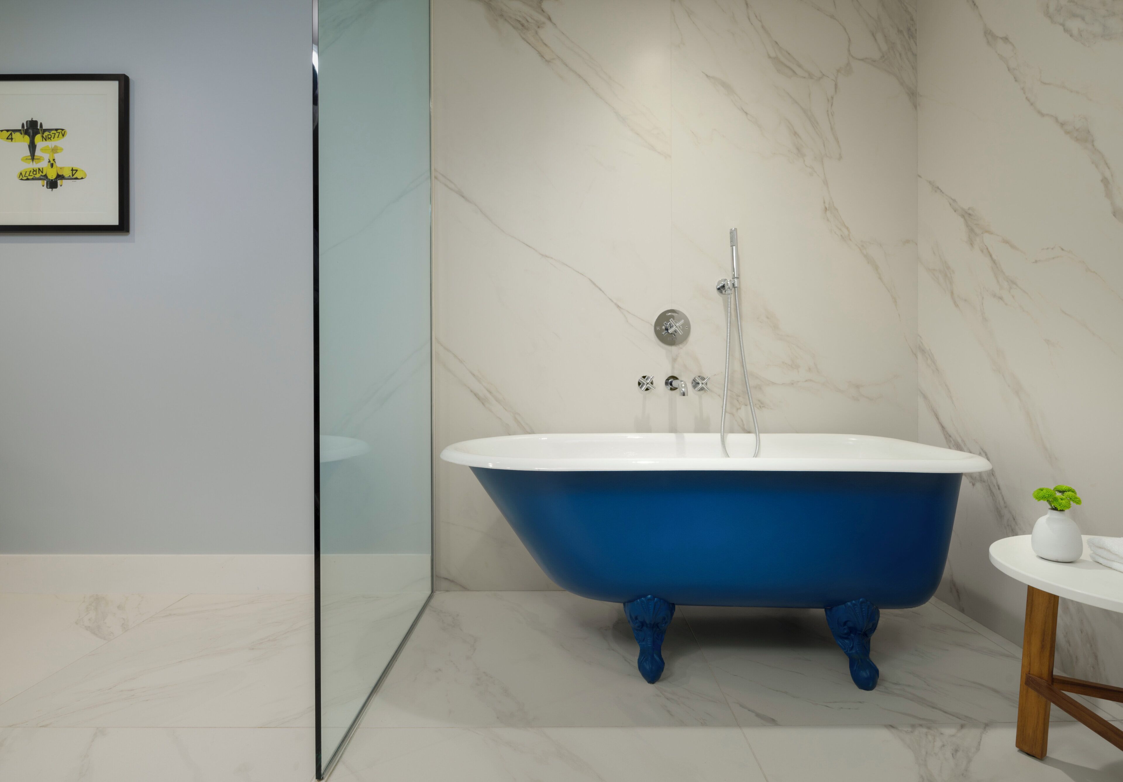 A modern bathroom with a blue clawfoot bathtub, a glass shower enclosure, and a marble wall.