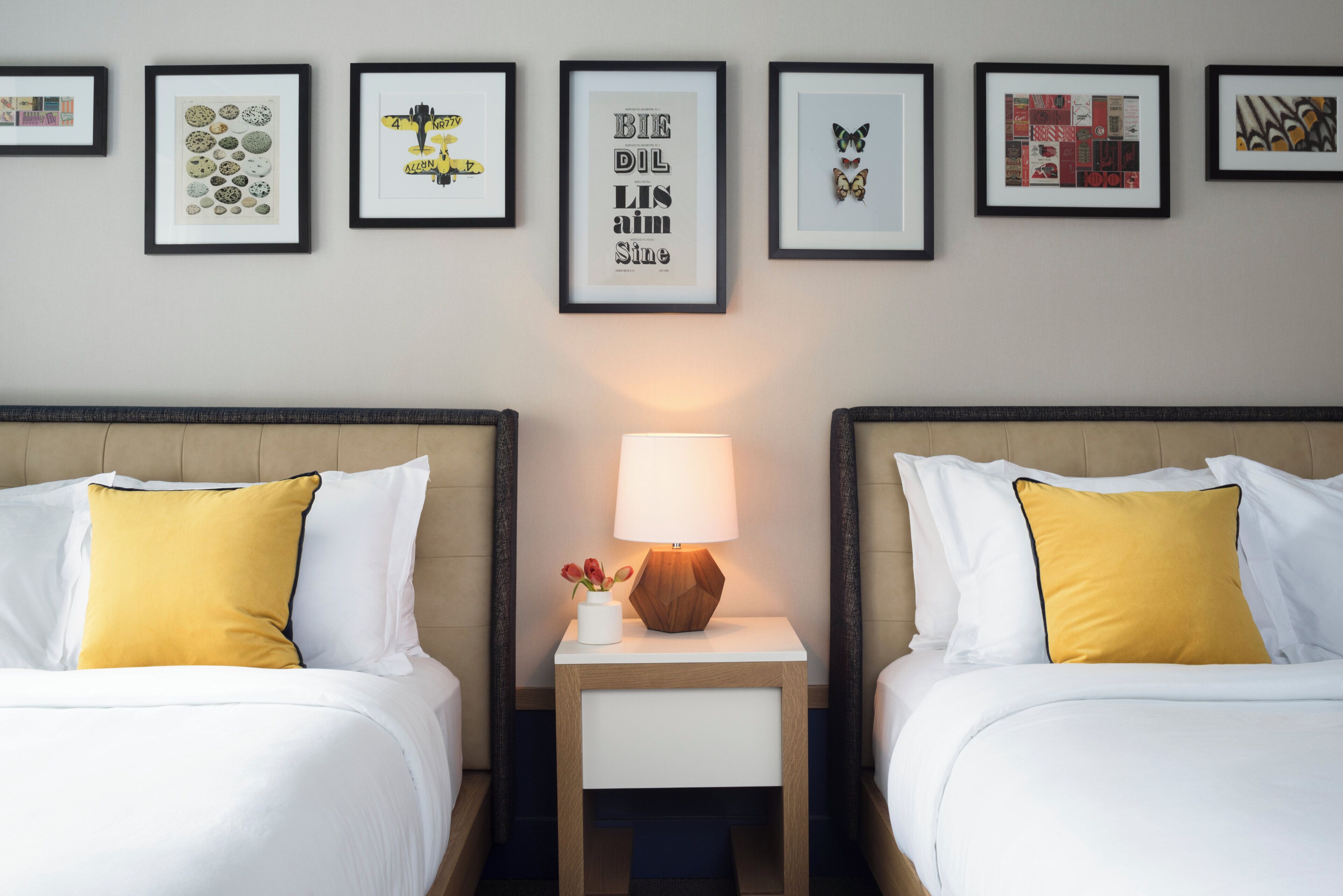 A hotel room with two beds, a nightstand with a lamp, and a wall decorated with framed pictures.
