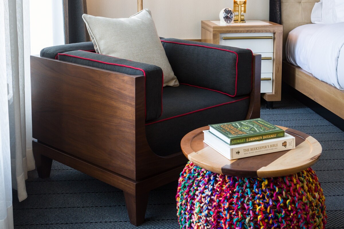 Een hotelkamer met een houten stoel, een klein nachtkastje met een lamp, een kleurrijke kruk en een bed met wit beddengoed.