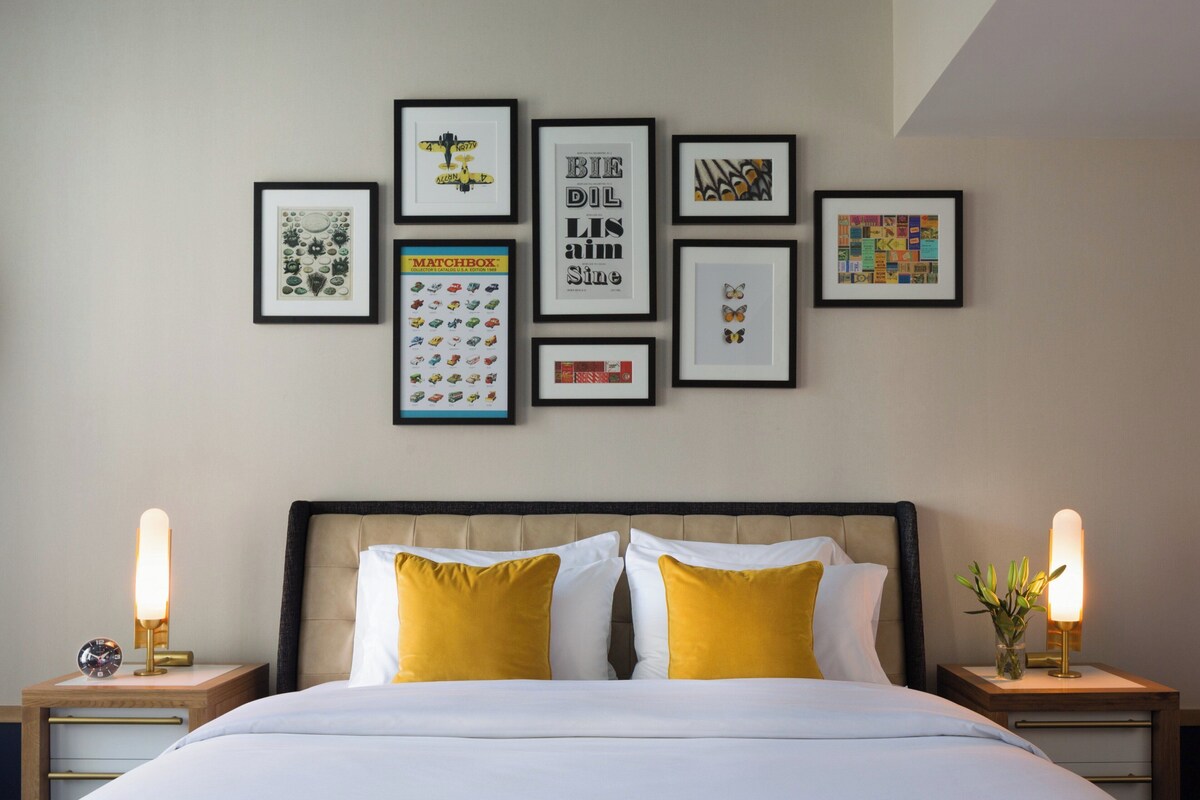 Een keurig opgemaakt slaapkamer met een bed, twee nachtkastjes met lampen, en een muur versierd met verschillende ingelijste kunstwerken.