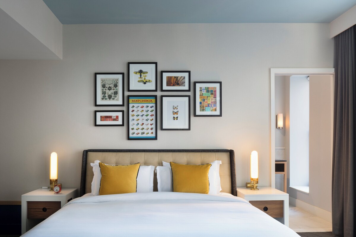 Een hotelkamer met een groot bed, twee nachtkastjes, een muur met ingelijste kunstwerken en een raam met gordijnen.