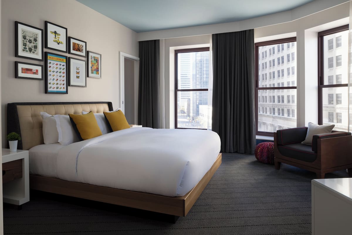A modern hotel room with a large bed, a desk, a chair, and a view of the city through large windows.