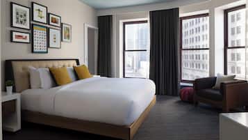 A modern hotel room with a large bed, a desk, a chair, and a view of the city through large windows.
