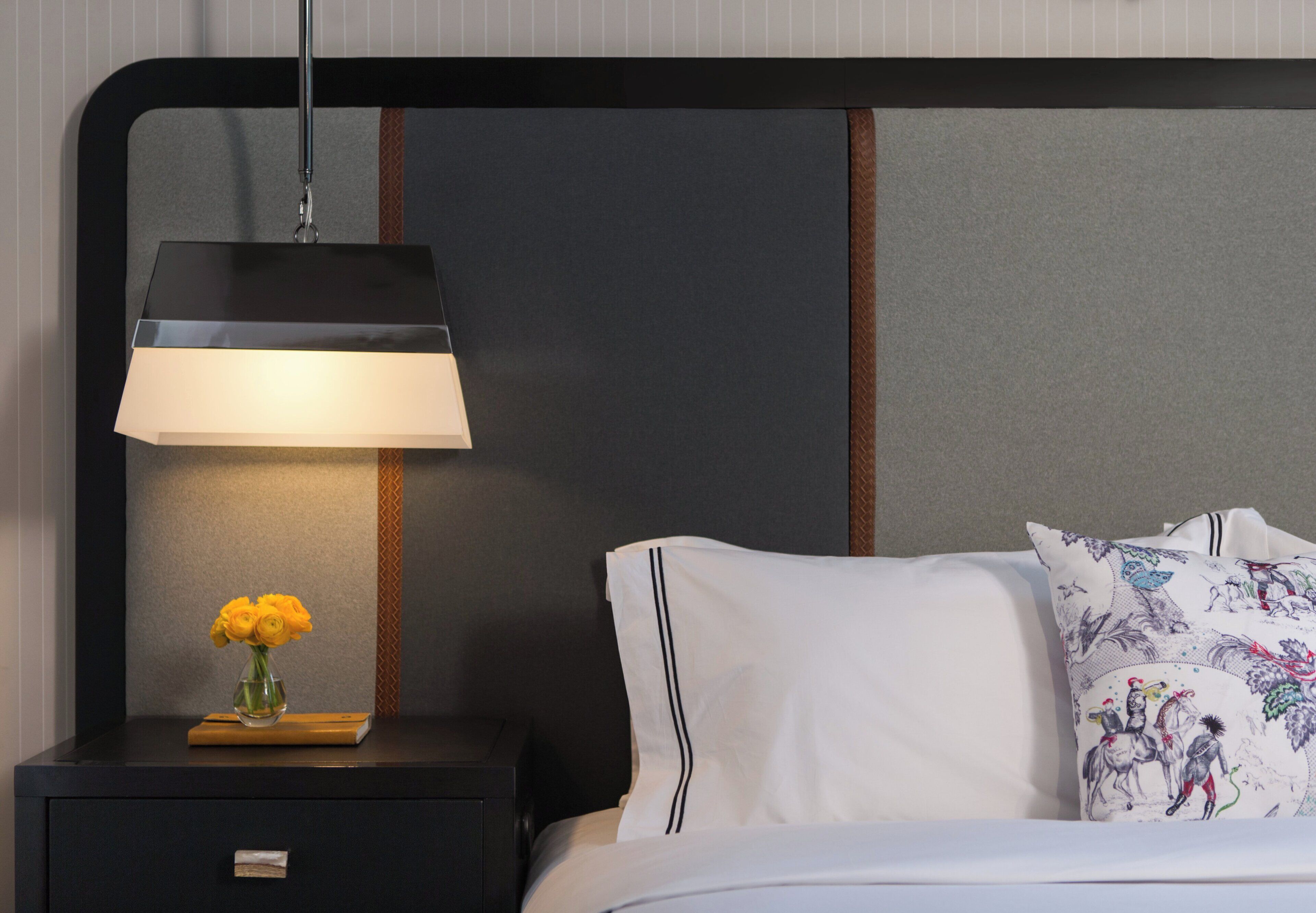A neatly made bed with white linens and decorative pillows, a bedside table with a lamp, and a vase with yellow flowers.
