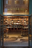 Bar com fachada de vidro, prateleiras repletas de garrafas e banquetas.