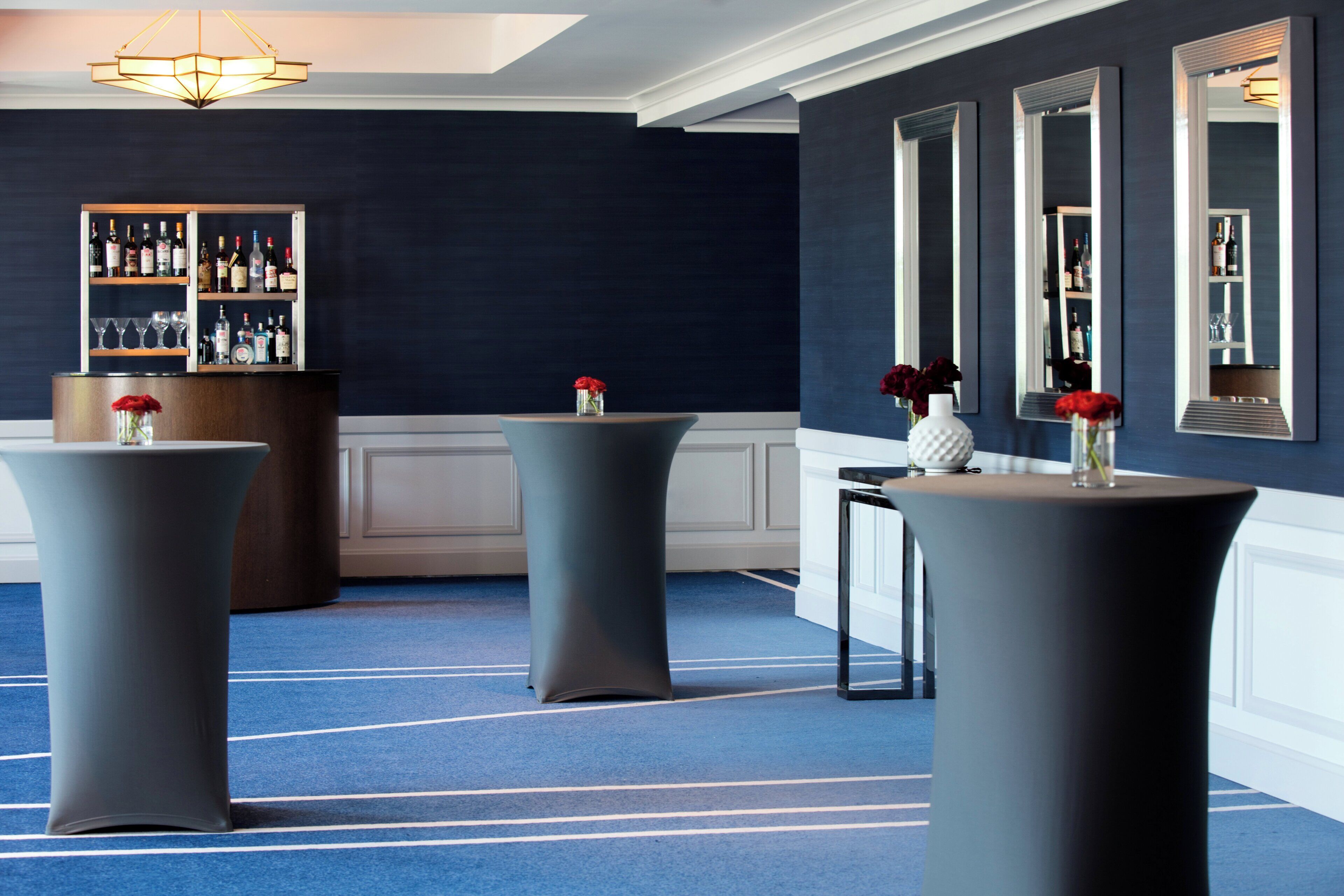 Un hall de hotel con tres mesas redondas, una zona de bar con estantes llenos de botellas y una pared azul oscuro con un espejo grande.