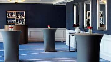 Un hall de hotel con tres mesas redondas, una zona de bar con estantes llenos de botellas y una pared azul oscuro con un espejo grande.