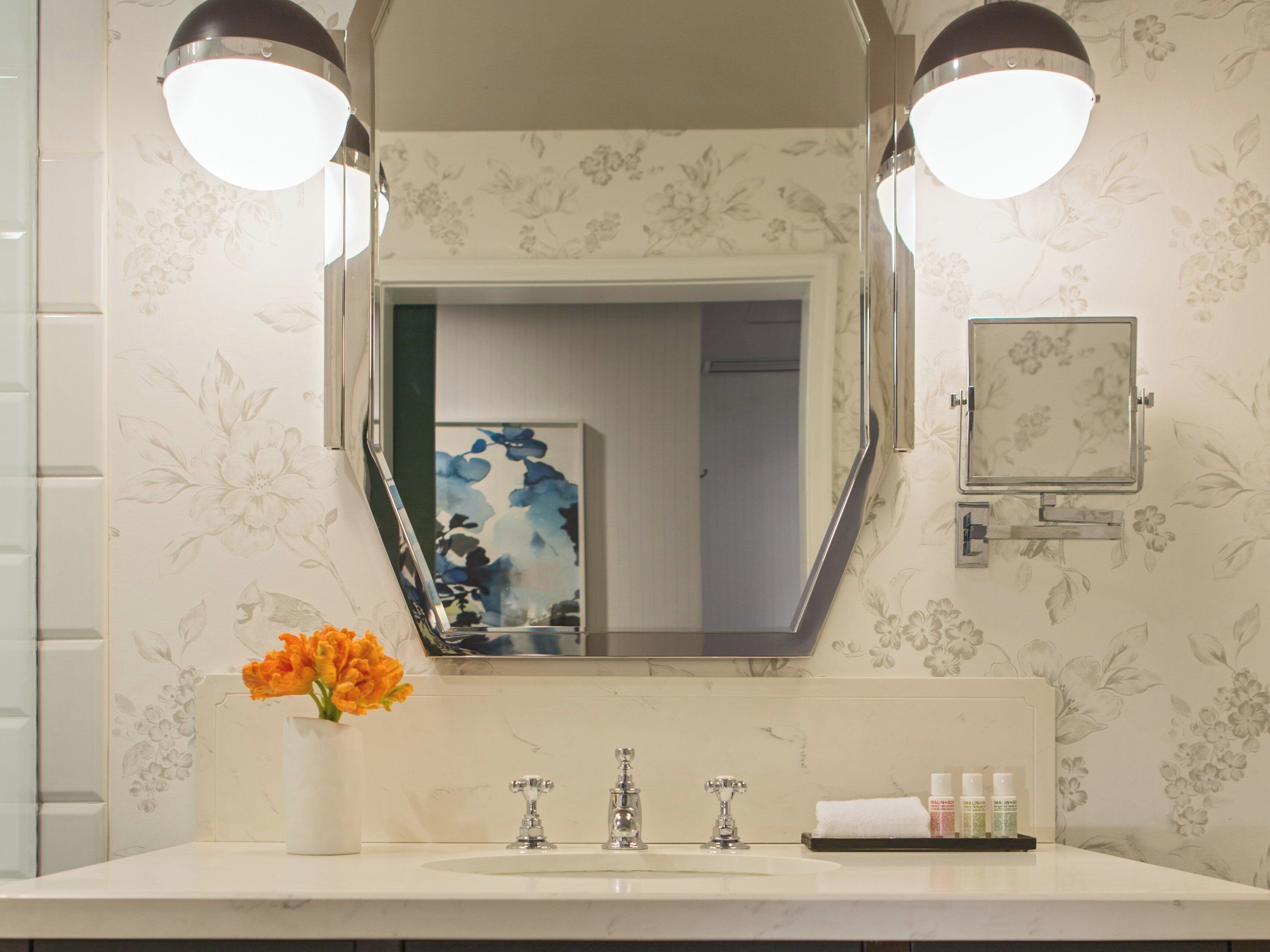 Un baño con un espejo grande, dos luces empotradas, un lavabo con tres grifos, un florero con flores y una pequeña foto enmarcada en la pared.