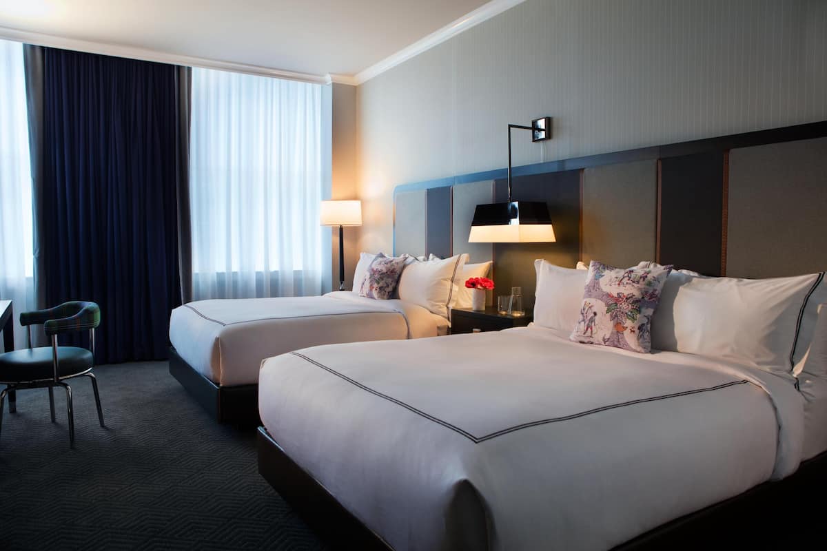 A hotel room with two beds, a desk, a chair, and a large window with curtains.
