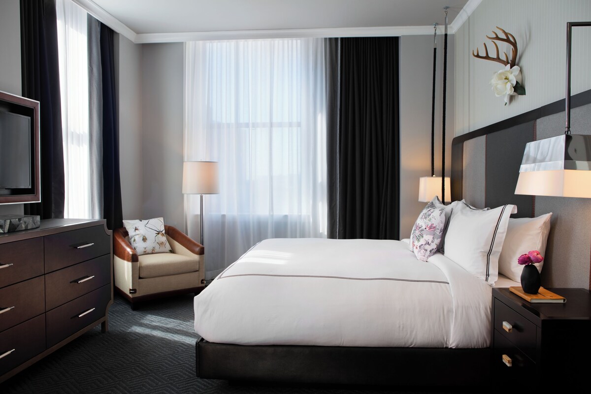 Habitación de hotel con una cama grande, televisión, escritorio con lámpara, una silla y ventana con cortinas.
