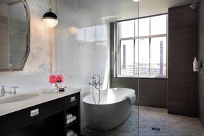 A modern bathroom with a freestanding bathtub, a large mirror, and a window offering a city view. - Kimpton Cardinal Hotel by IHG (Winston-Salem)