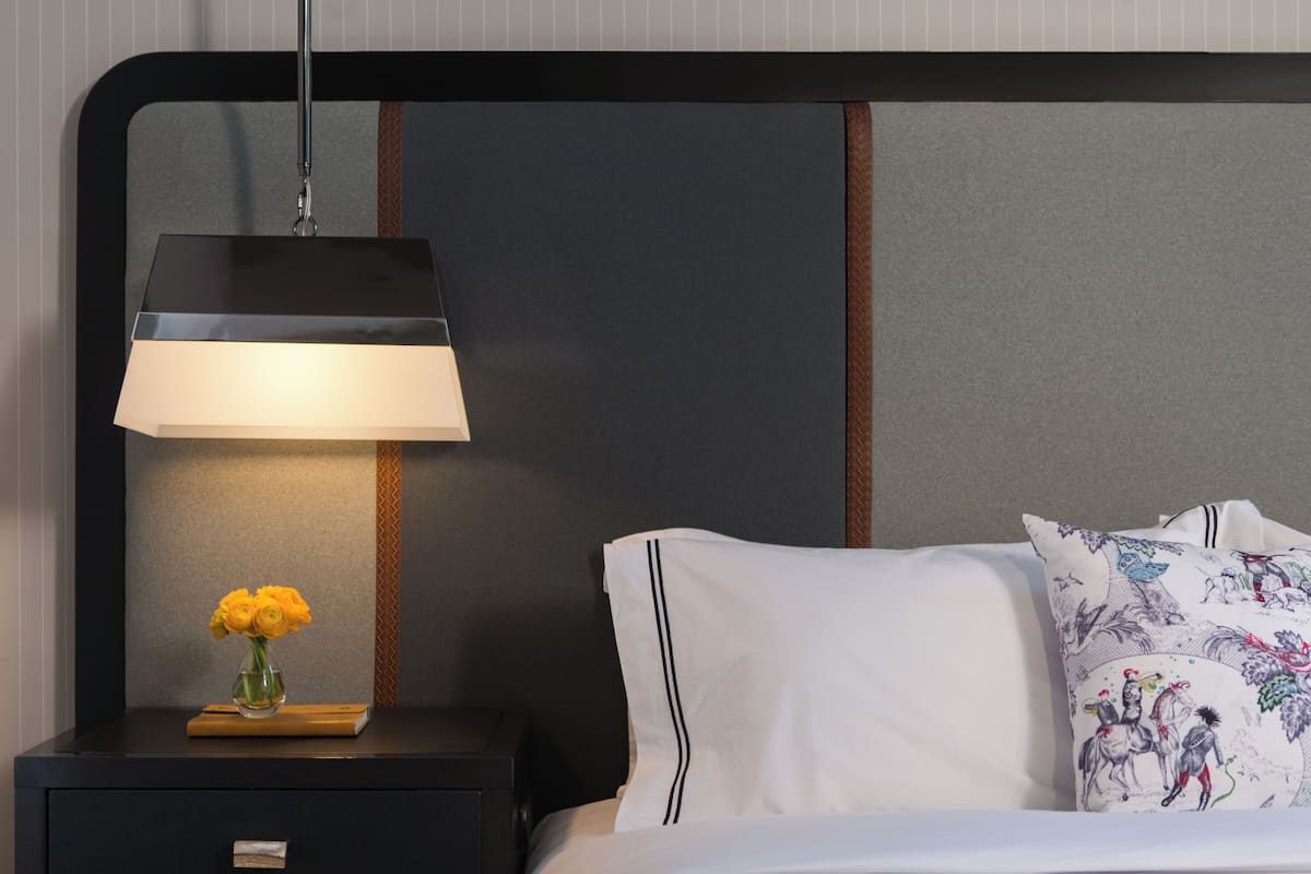 Ein ordentlich bezogenes Bett mit weißer Bettwäsche und dekorativen Kissen, ein Nachttisch mit Lampe und Blumenvase sowie ein modernes Kopfstück mit Streifenmuster.