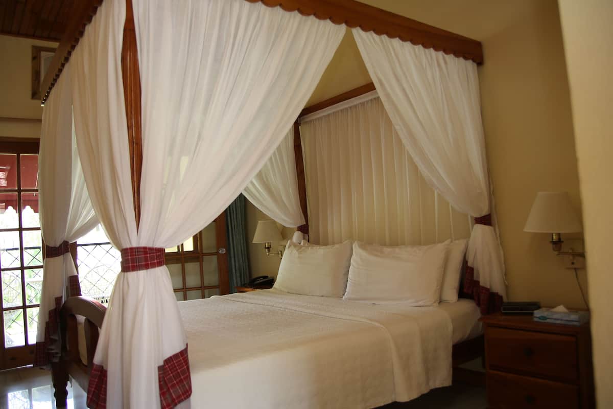A four-poster canopy bed with white linens and red accents, a bedside table with a lamp, and a window with curtains.