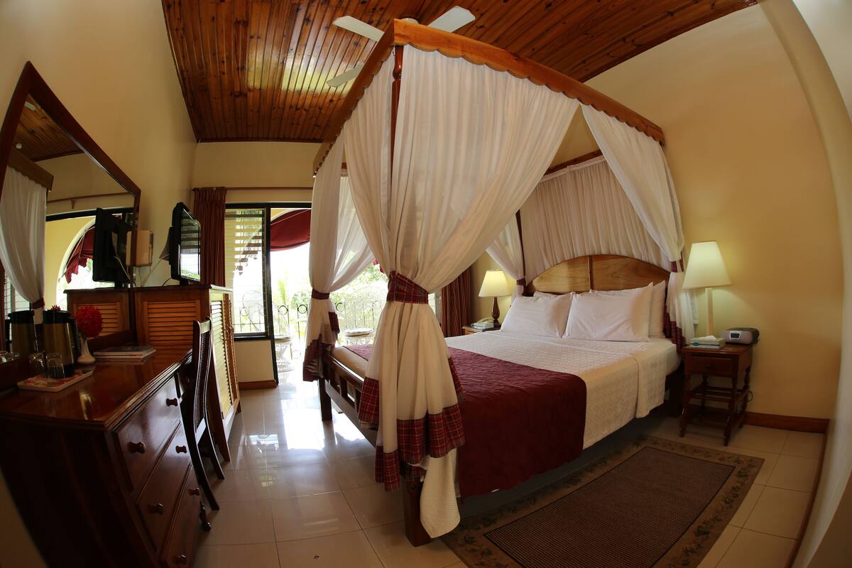 A four-poster canopy bed with a red bedspread, a wooden dresser with drawers, a mirror, and a small table with a lamp.
