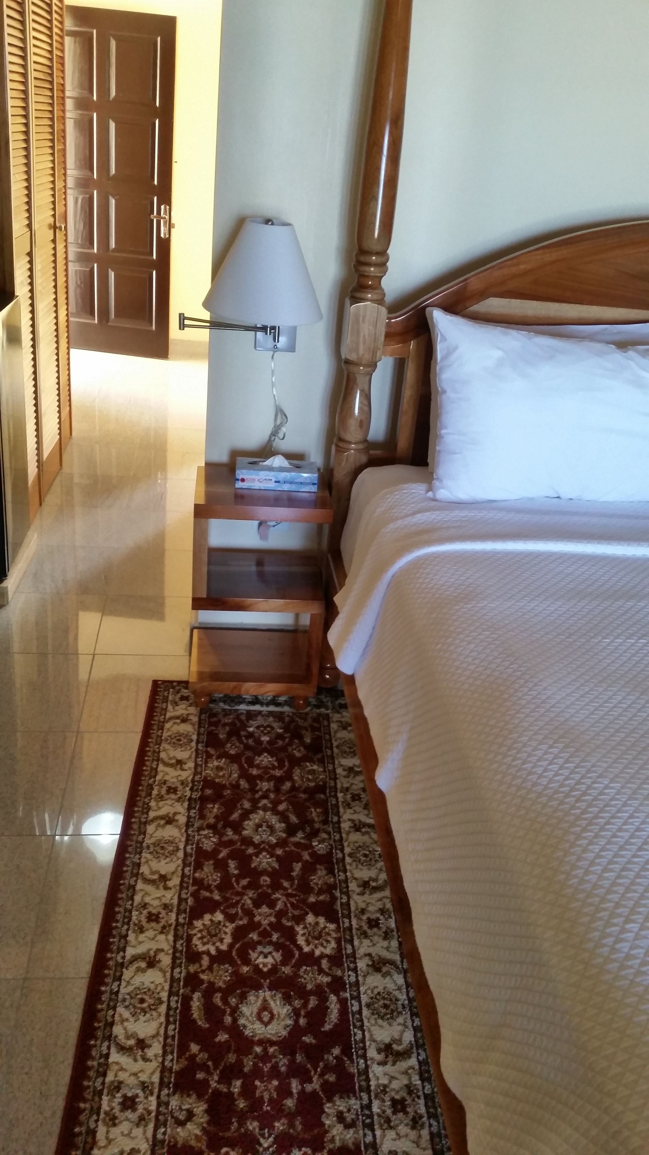 A hotel room with a wooden bed, a nightstand with a lamp, a patterned rug, and a door leading to another room.