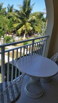 Varanda com uma mesa redonda e duas cadeiras, com vista para um jardim com palmeiras e uma cerca.