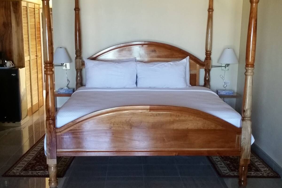A four-poster bed with a wooden frame and white bedding, a ceiling fan, and a tiled floor.