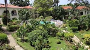 Un jardin luxuriant avec une piscine, des palmiers et un sentier menant à un bâtiment surmonté d’un toit recouvert de tuiles rouges.