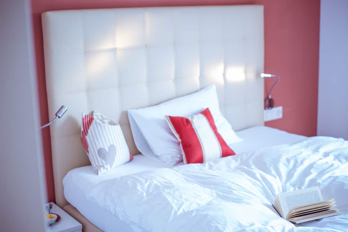 Una cama bien hecha con ropa blanca y almohadas rojas y blancas, una lámpara de noche y un libro sobre la cama.