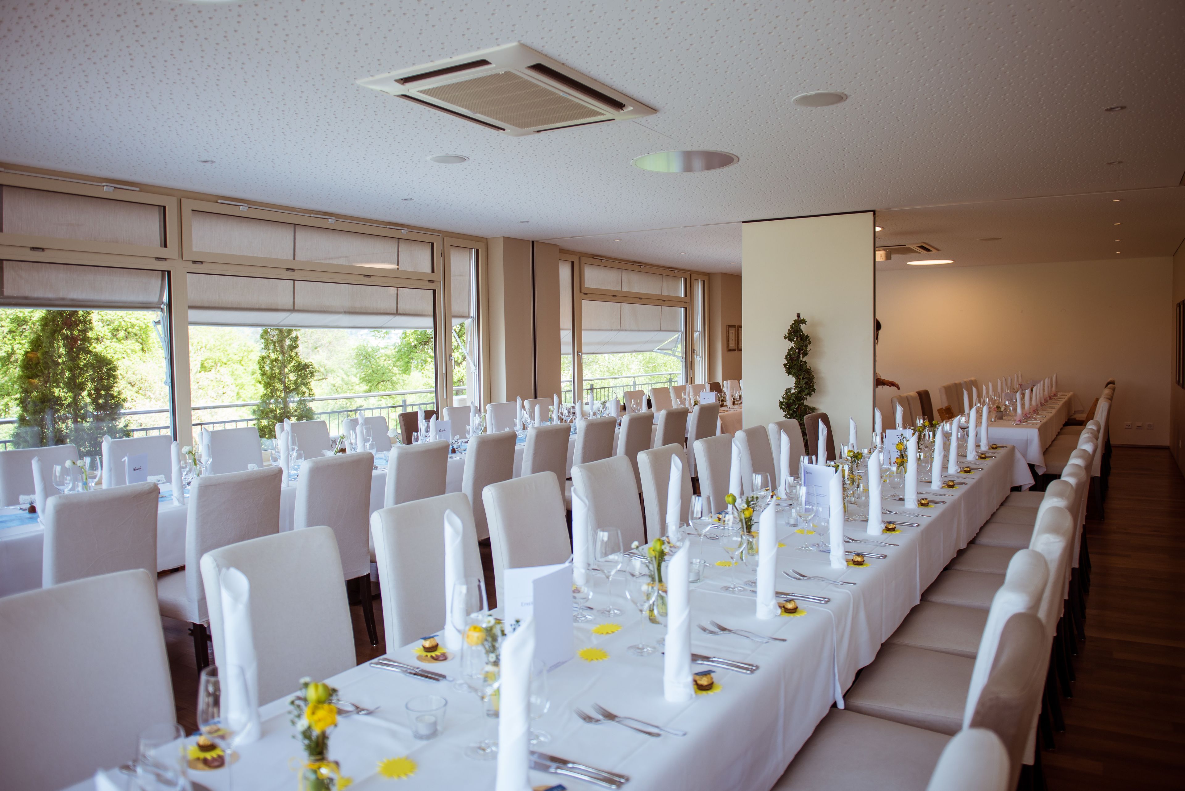A banquet hall with long tables set for a formal event, large windows, and a ceiling fan.
