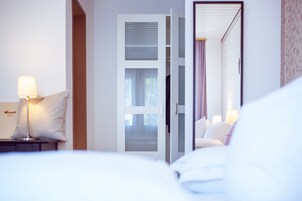 Habitación de hotel con cama, mesita de noche con lámpara, y puerta abierta que da a otra habitación.