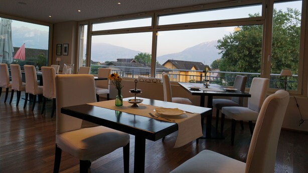 A dining area with a table set for two, large windows offering a view of mountains and houses, and a row of white chairs. - Hotel Restaurant Weinstube (Eschen)